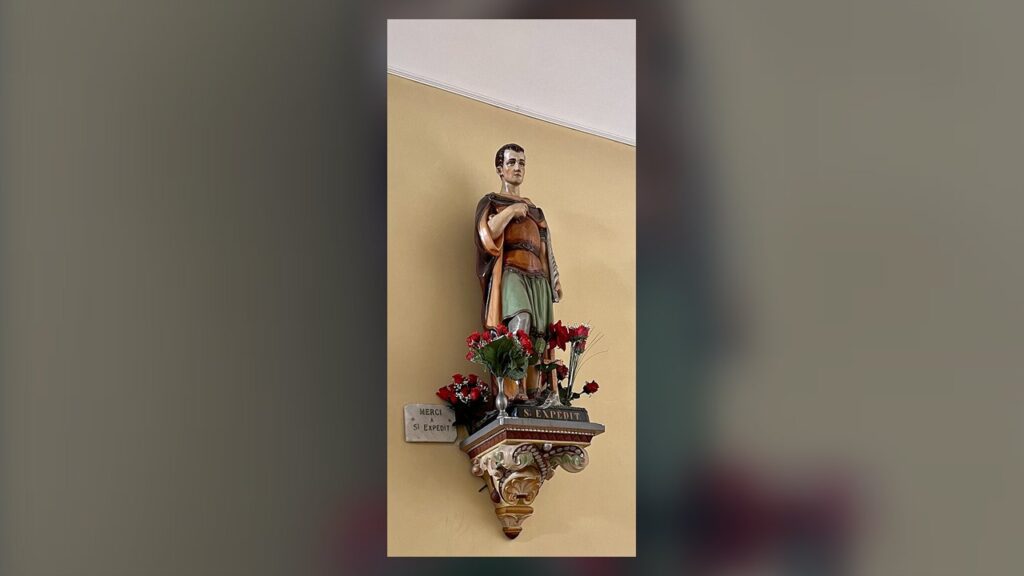 Estátua de Santo Expedito com flores e placa de agradecimento, sinal da devoção popular ao santo