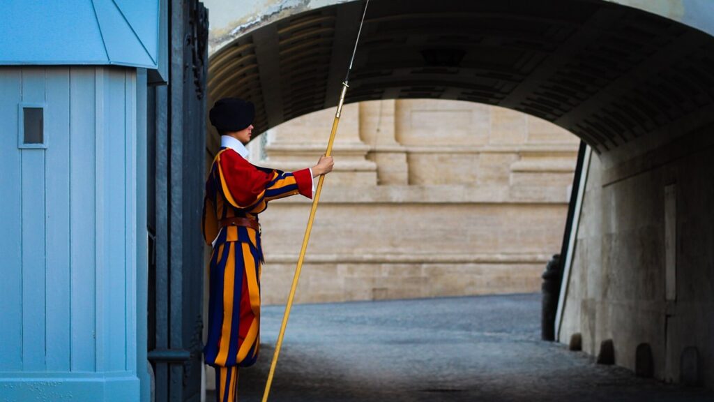 Guarda suíço com alabarda e uniforme de gala azul, vermelho e amarelo numa entrada do Vaticano