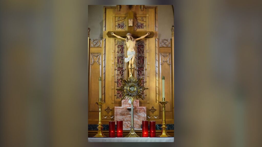 Hóstia exposta no ostensório sobre o altar, entre velas, diante do crucifixo, durante a adoração eucarística