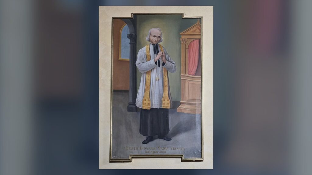 São João Maria Vianney de batina, sobrepeliz e estola ao lado do confessionário, modelo do padre diocesano
